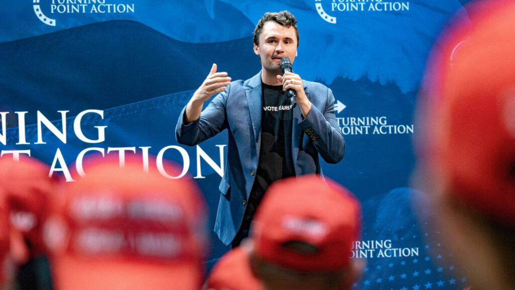 Charlie Kirk Mobilizes the Youth Vote at Turning Point Action – Tumisang Bogwasi Charlie Kirk speaking into a microphone at a Turning Point Action event, standing in front of a blue backdrop with red-hatted attendees in the foreground.