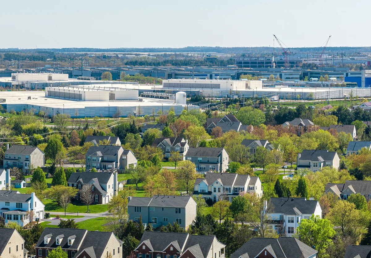Data Center Alley Meets the Suburbs — Loudoun County, VA – Tumisang Bogwasi Suburban homes and trees in the foreground with large white data-center buildings, power lines, and cranes in the background in Loudoun County, Virginia.
