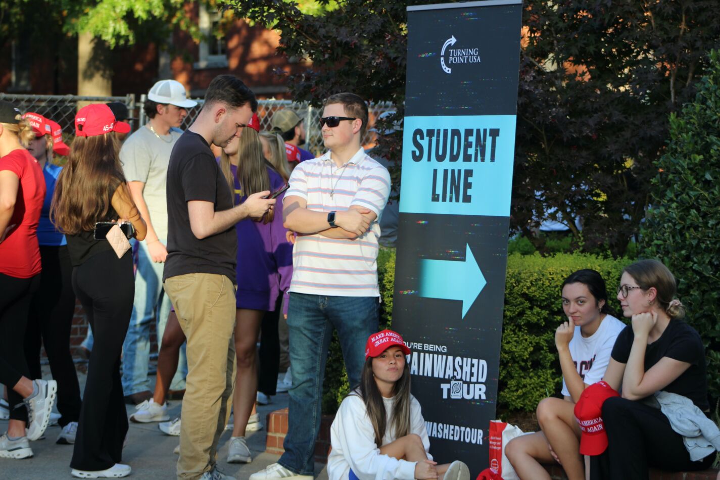 Students Line Up for Turning Point USA’s “Brainwashed Tour” – Tumisang Bogwasi Students gathered near a “Student Line” sign at a Turning Point USA event, with some wearing red MAGA hats and others sitting near the banner reading “Culture Being Brainwashed Tour.”