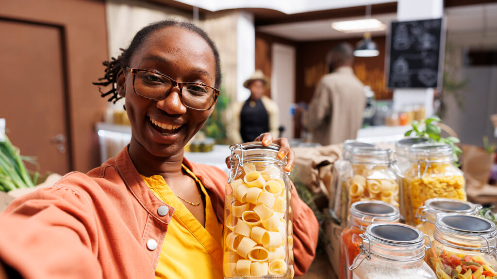 Smiling person in glasses holds a jar of large pasta shapes while taking a selfie in a bright refill shop with rows of glass jars and plants in the background.