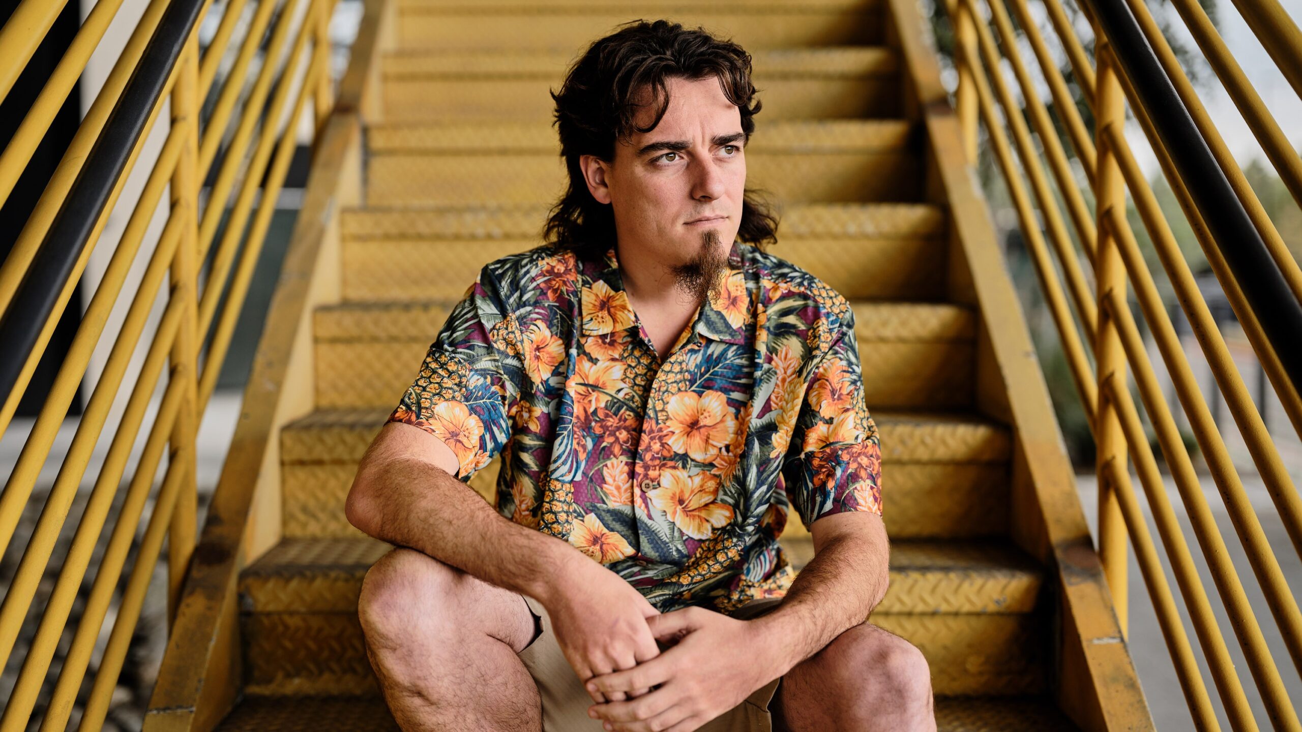 Palmer Luckey Rebel Technologist on the Warpath – Tumisang Bogwasi Palmer Luckey, founder of Anduril, seated on yellow metal stairs wearing a colorful floral shirt, looking off thoughtfully.
