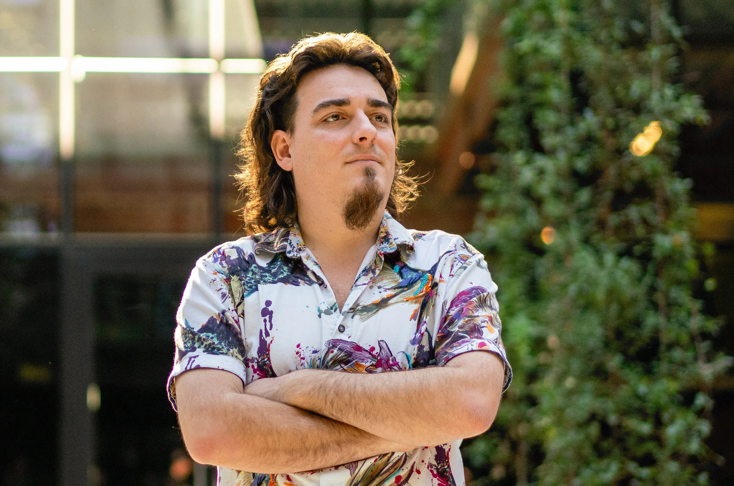Palmer Luckey The Maverick Behind Anduril – Tumisang Bogwasi Palmer Luckey, founder of Anduril, standing confidently with arms crossed in a colorful shirt, outdoors with greenery in the background.
