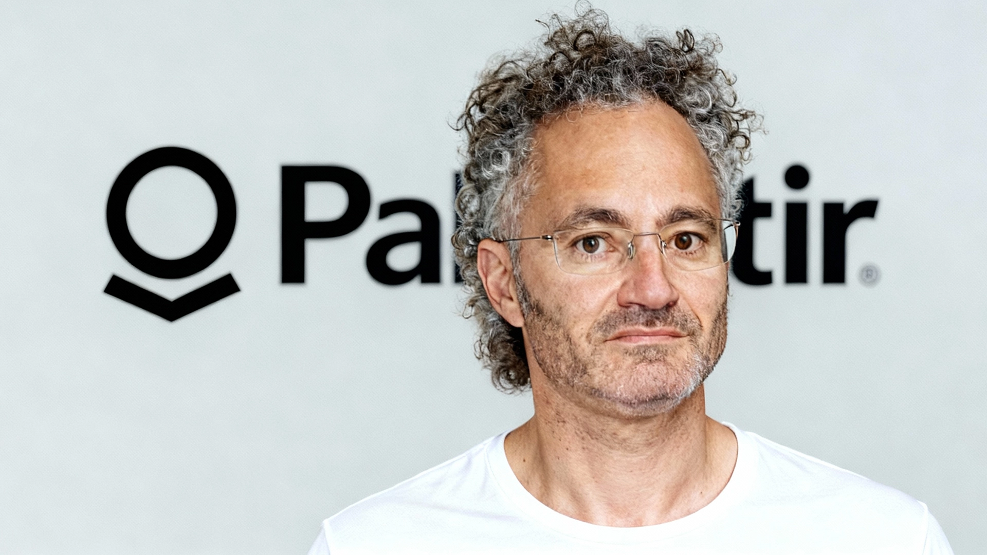 Alex Karp wearing glasses and a white shirt standing in front of the Palantir logo