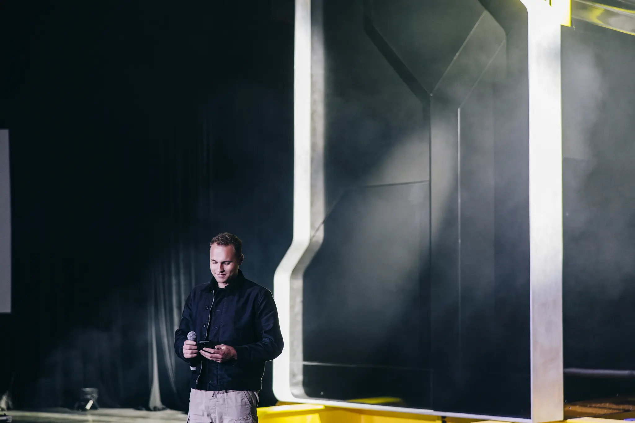 Isaiah Taylor Unveils the Future of Energy – Tumisang Bogwasi Isaiah Taylor holding a microphone and smartphone on stage in front of Valar Atomics reactor structure, preparing to speak.