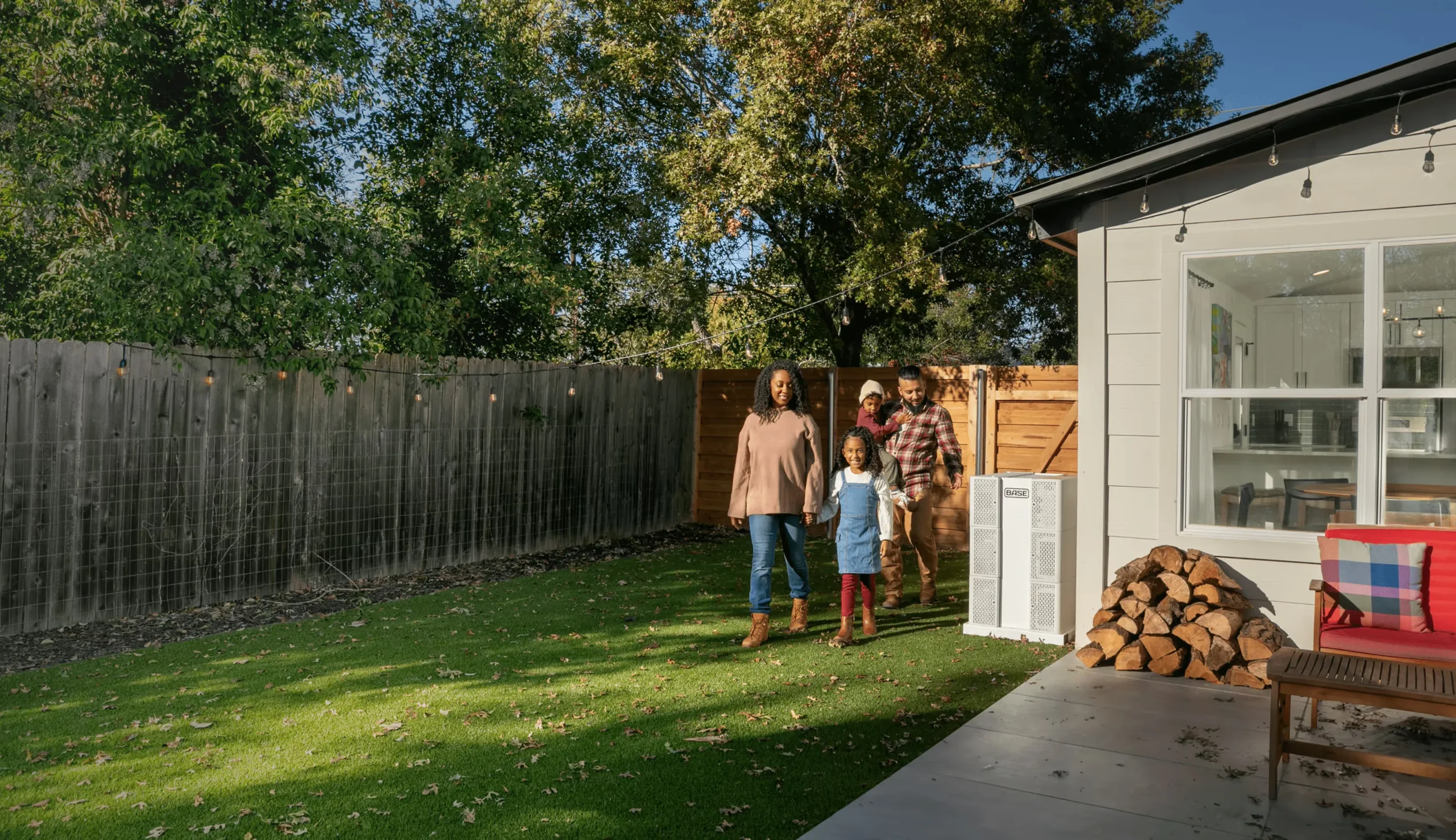 Base Home Energy Made for Everyday Life – Tumisang Bogwasi A family walks across a backyard toward a modern home, with a Base home energy system installed beside the house, surrounded by trees and warm afternoon light.