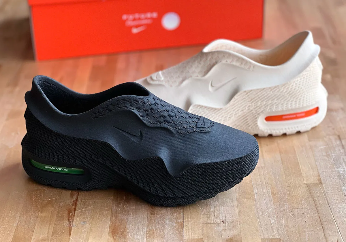 Black and bone-colored Nike Air Max 1000 shoes displayed on a wooden surface with an orange Nike box in the background.