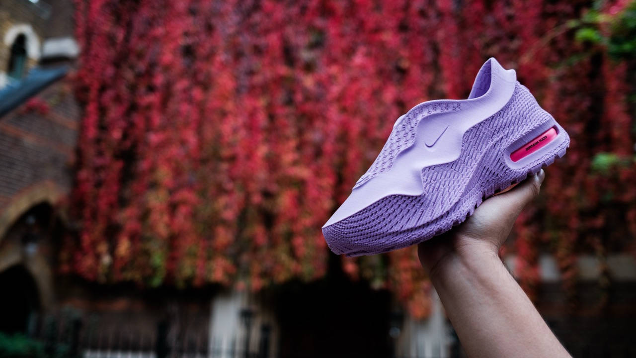 A hand holding a lavender Nike Air Max 1000 sneaker with a textured upper and visible pink Air unit, against a blurred red foliage background.