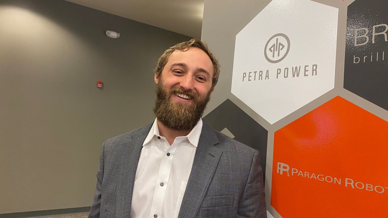 Aaron Goodman, co-founder of Petra Power, smiling in front of a Petra Power logo wall at the company's headquarters.