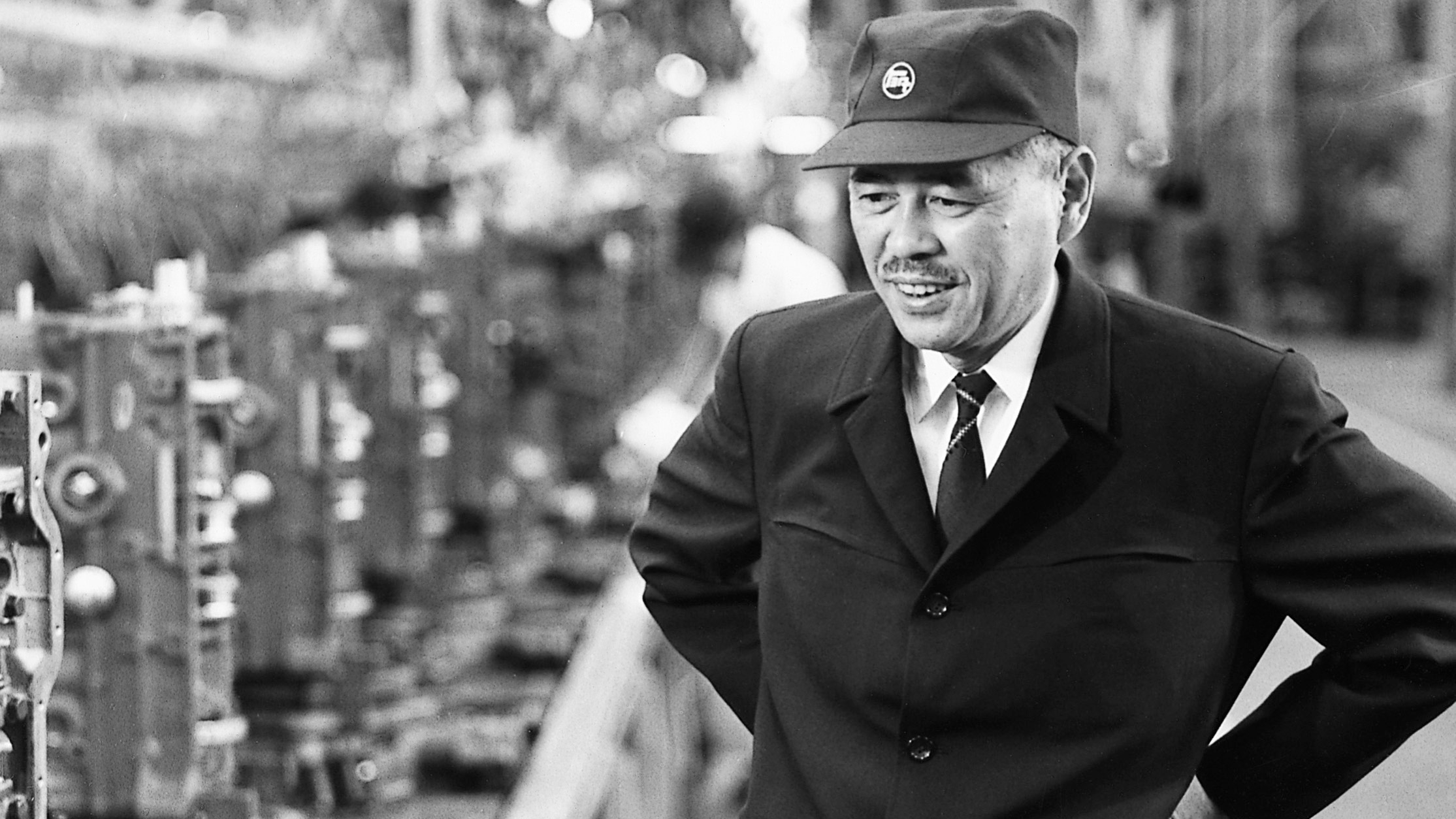 Taiichi Ohno smiling and standing on a factory floor in uniform and cap, surrounded by industrial machinery