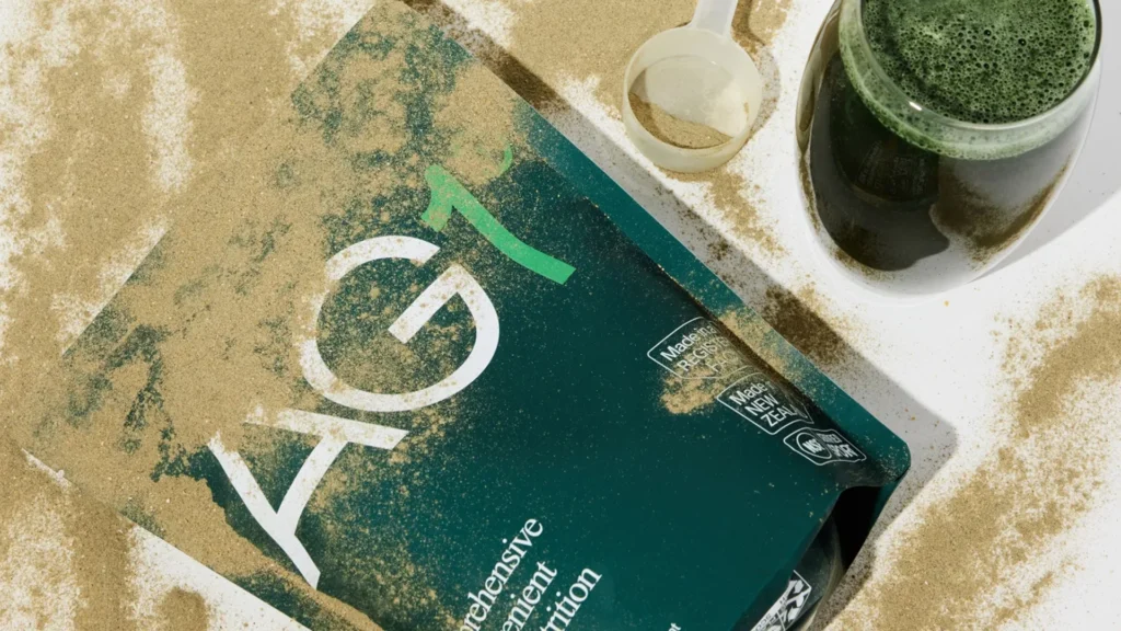 AG1 Greens Powder and Prepared Drink – Tumisang Bogwasi Open AG1 greens powder pouch with powder spilled around it, next to a scoop and a glass of green drink on a white background.