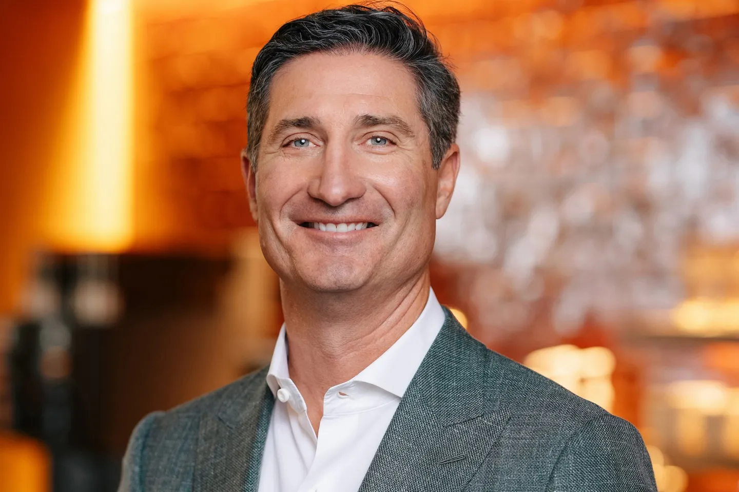 Portrait of Brian Niccol, CEO of Starbucks, smiling and wearing a gray blazer and white shirt against a warmly lit background.