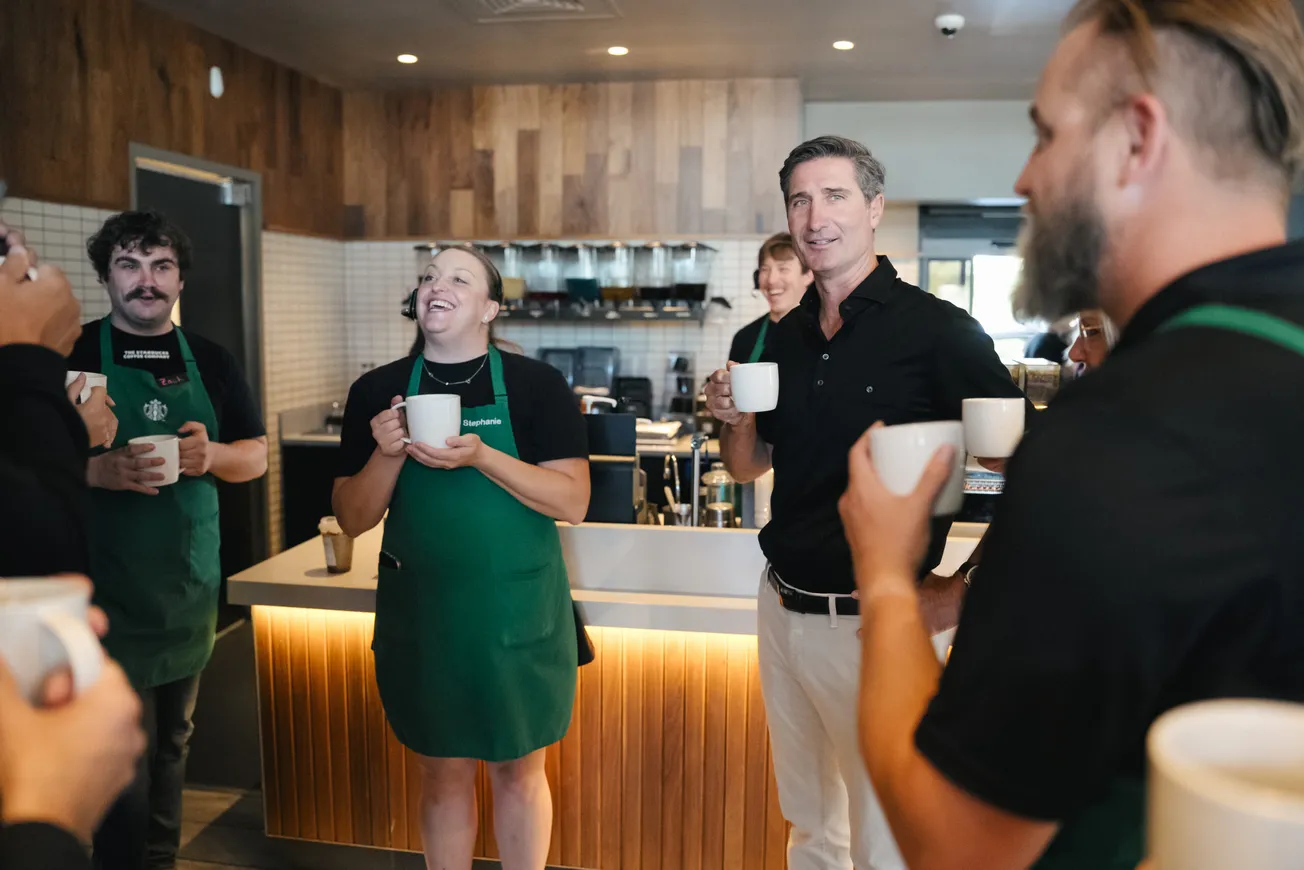 Brian Niccol, CEO of Starbucks, standing inside a Starbucks café with employees wearing green aprons, all holding coffee mugs and smiling.