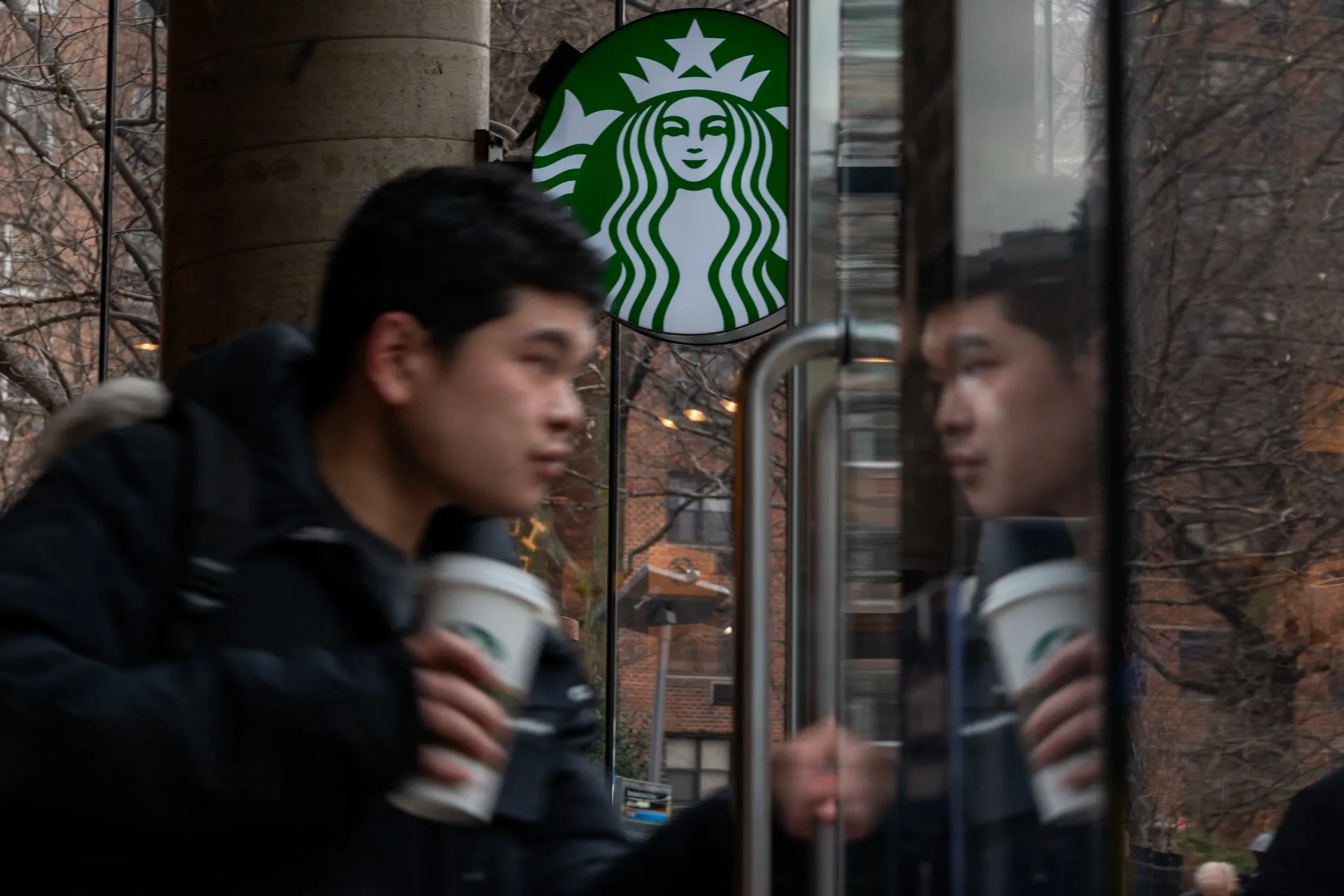 A man holding a Starbucks takeaway cup opens a glass door to enter a Starbucks store, with the green Starbucks logo visible above him.