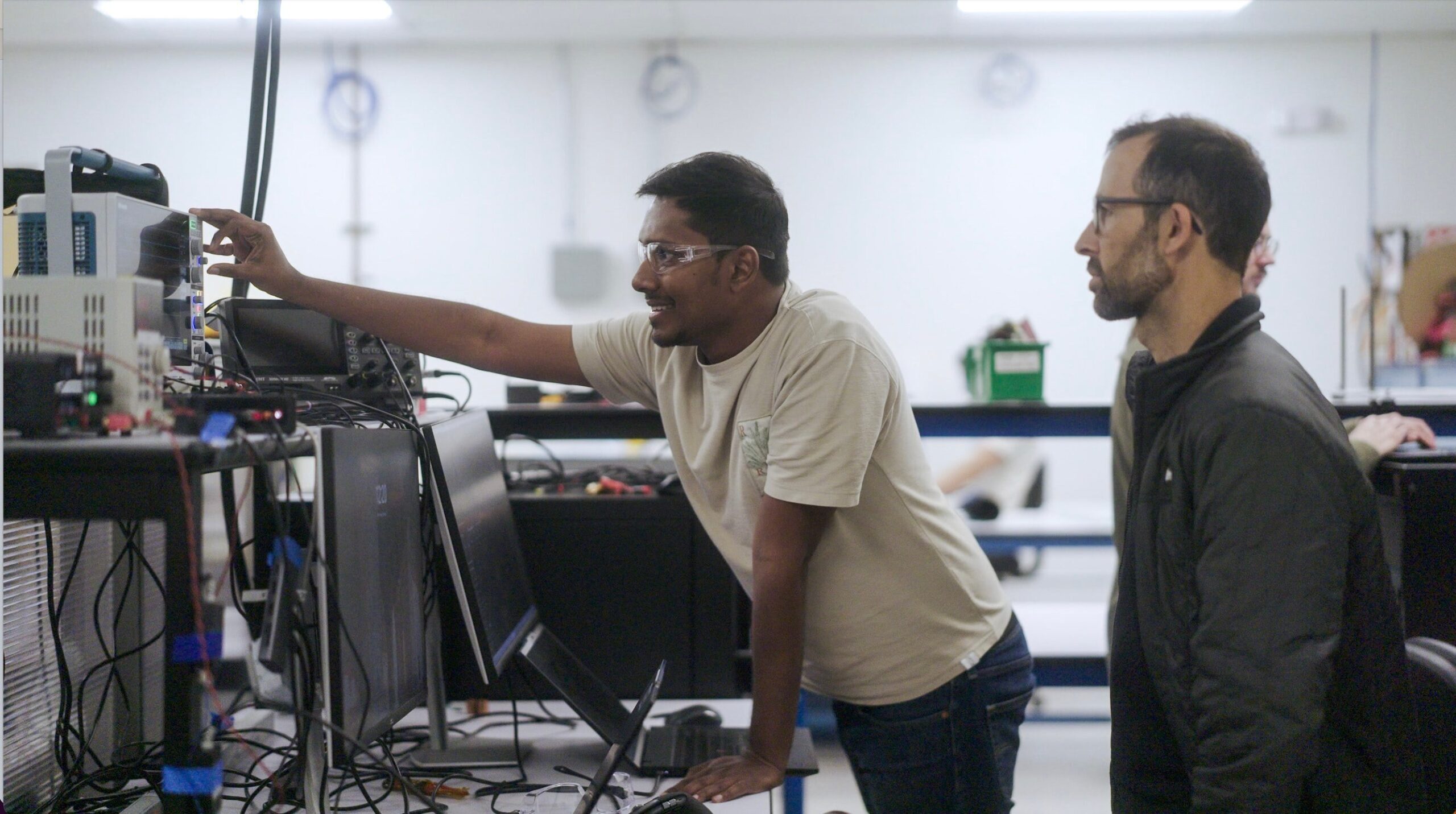 Drew Baglino standing beside an engineer wearing safety glasses as they adjust and monitor electronic testing equipment in a laboratory setting.