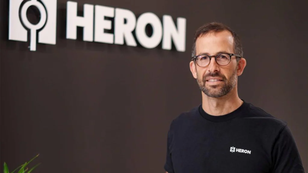 Drew Baglino wearing glasses and a black Heron T-shirt, standing in front of a wall with the Heron company logo.