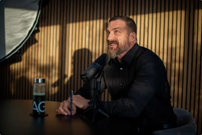 Podcast Recording Session with AG1 Branding – Tumisang Bogwasi Bearded man in a black shirt speaking into a podcast microphone at a desk, with an AG1-branded water bottle nearby and wooden paneling in the background.