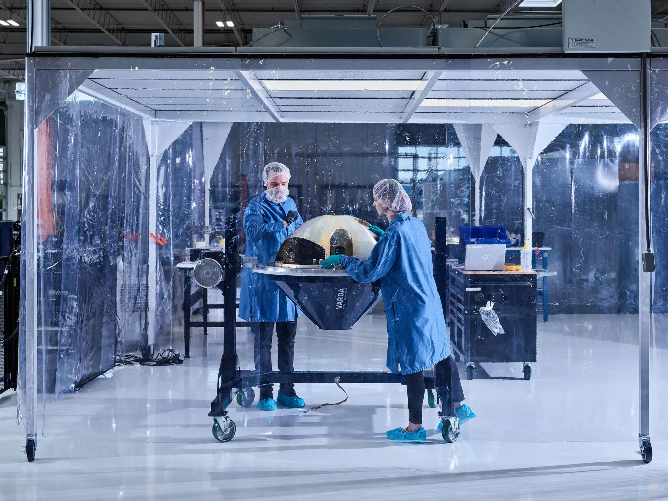 Two technicians in cleanroom attire inspect a Varda reentry capsule inside a transparent containment enclosure in a laboratory.