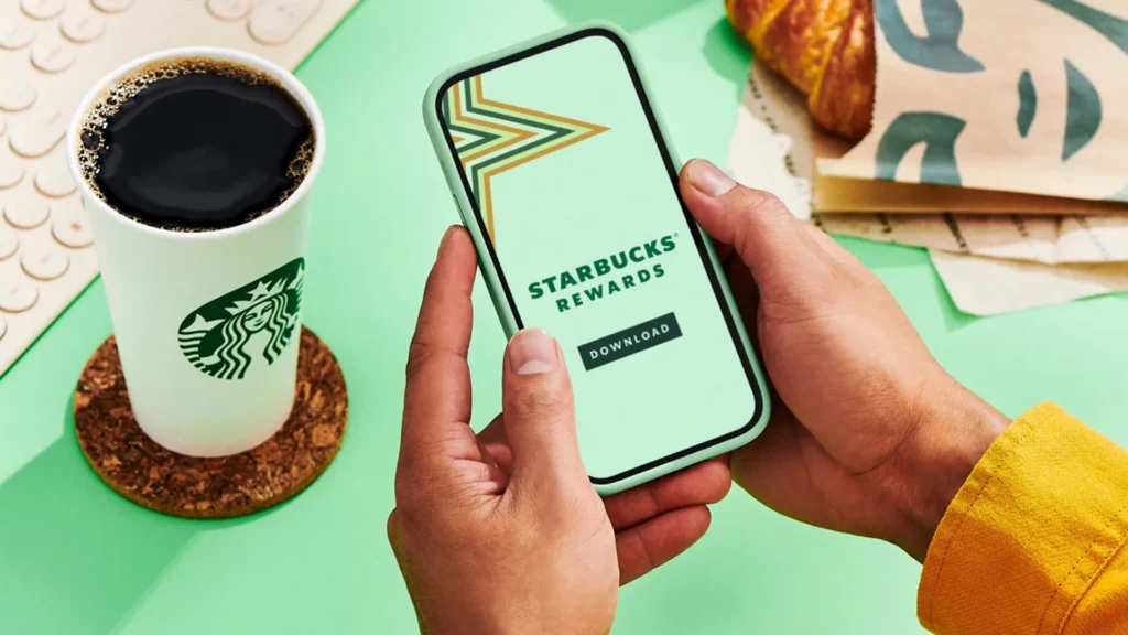 Hands holding a smartphone showing the Starbucks Rewards app download screen, next to a Starbucks coffee cup and a croissant on a green table.