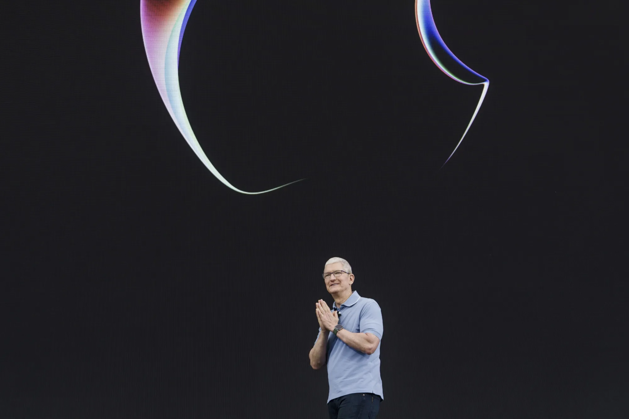 Tim Cook at apple event – Tumisang Bogwasi Tim Cook standing on a dark stage during an Apple keynote, hands clasped, with abstract colorful light graphics above him.
