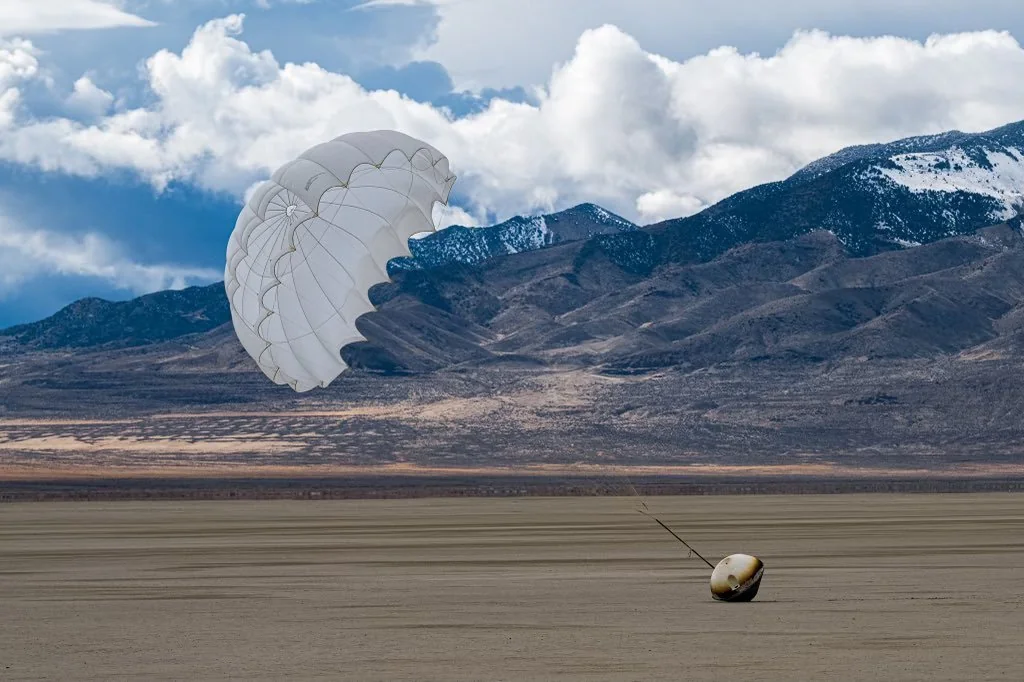 Varda reentry capsule landing in a desert under a deployed parachute, with mountains and clouds in the background.