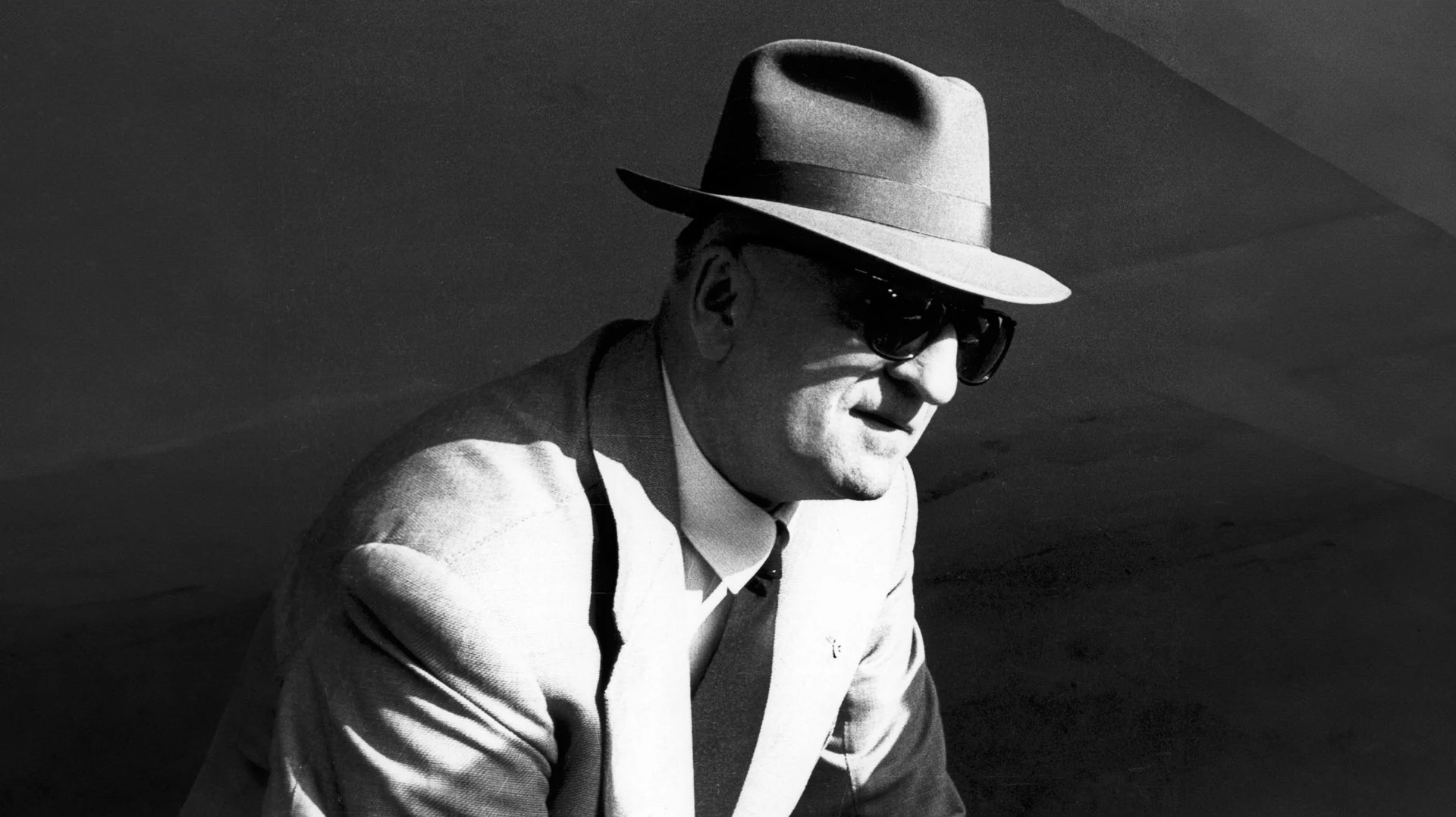 Enzo Ferrari Portrait in Black and White – Tumisang Bogwasi Black-and-white photograph of Enzo Ferrari in a suit and tie, wearing a fedora and dark sunglasses, seated in profile.