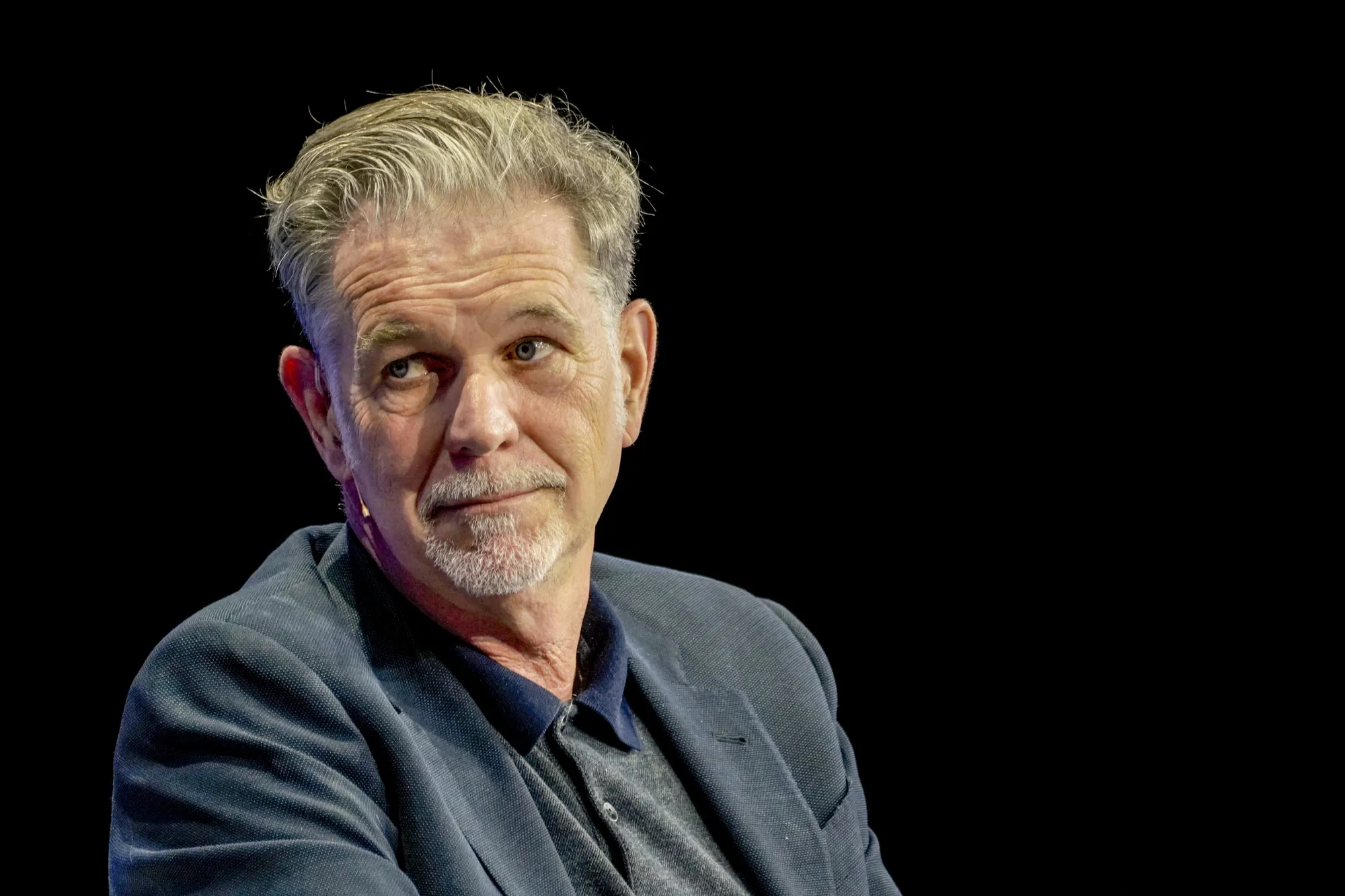 Reed Hastings Portrait at Public Event – Tumisang Bogwasi Reed Hastings looking to the side while seated on stage against a dark background.