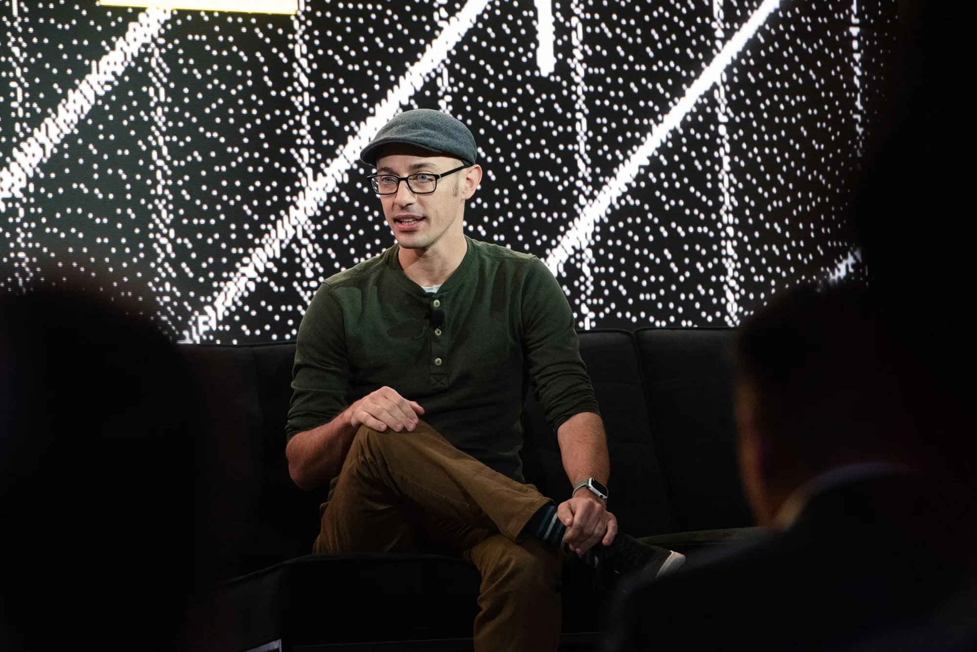Shopify CEO Tobi Lütke sitting on a stage during a tech talk, wearing glasses and a cap with a digital display backdrop.