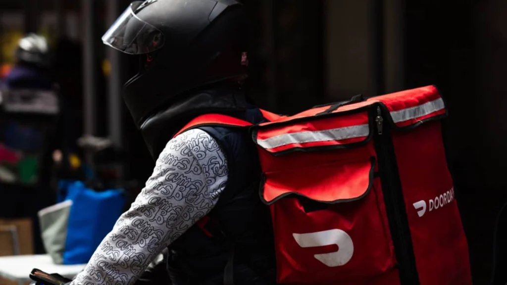 DoorDash Courier with Branded Delivery Bag – Tumisang Bogwasi A DoorDash delivery rider wearing a black helmet and carrying a red DoorDash insulated backpack while seated on a bicycle or scooter.