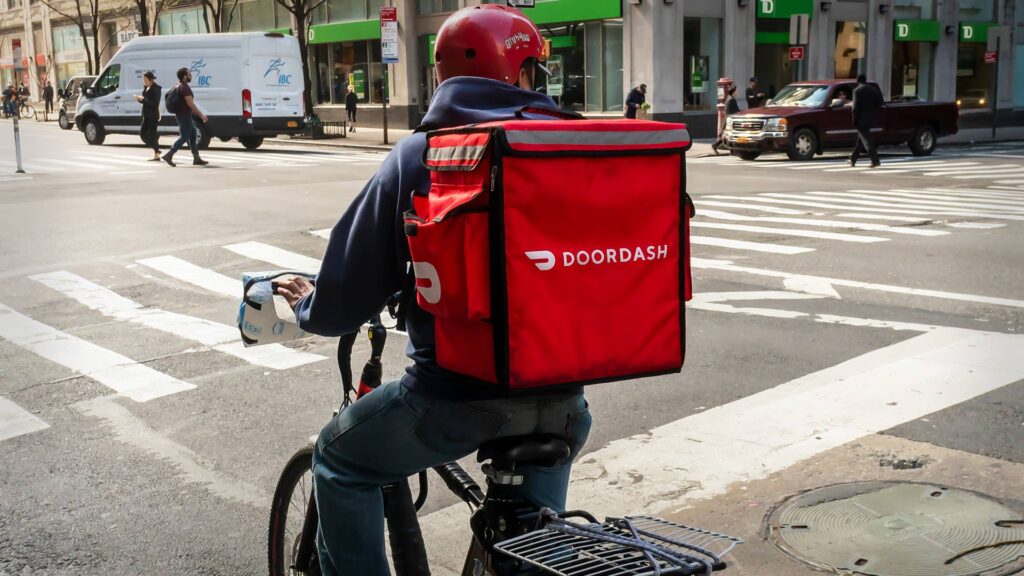 DoorDash Delivery Rider in an Urban Street Scene – Tumisang Bogwasi A DoorDash delivery rider wearing a red helmet and carrying a red DoorDash delivery bag while seated on a bicycle at a city intersection.