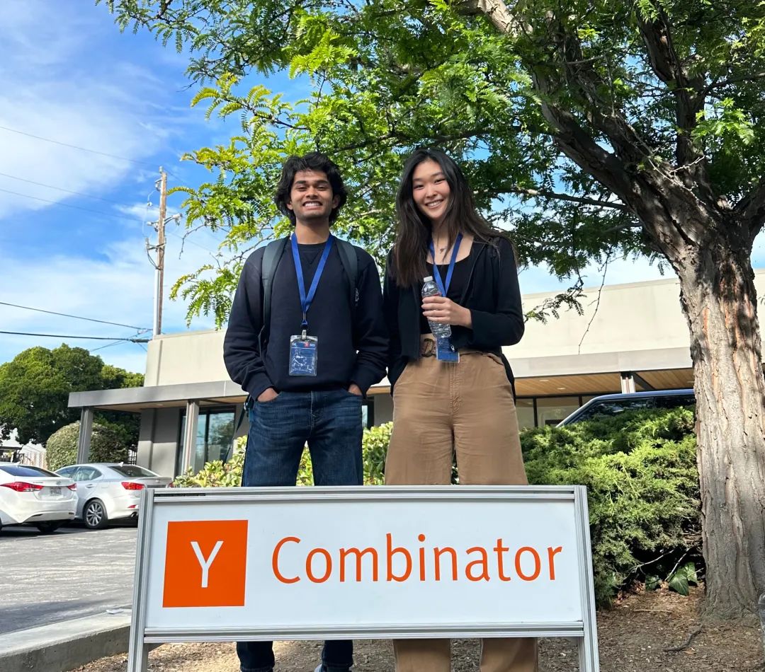 Jessica Wu and Neil Deshmukh standing outdoors behind a Y Combinator sign, wearing badges and smiling.