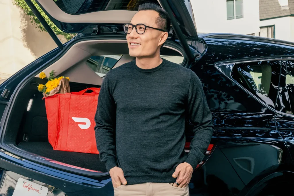 Tony Xu by a DoorDash Delivery Vehicle – Tumisang Bogwasi Tony Xu standing beside an open car trunk with a red DoorDash delivery bag inside.