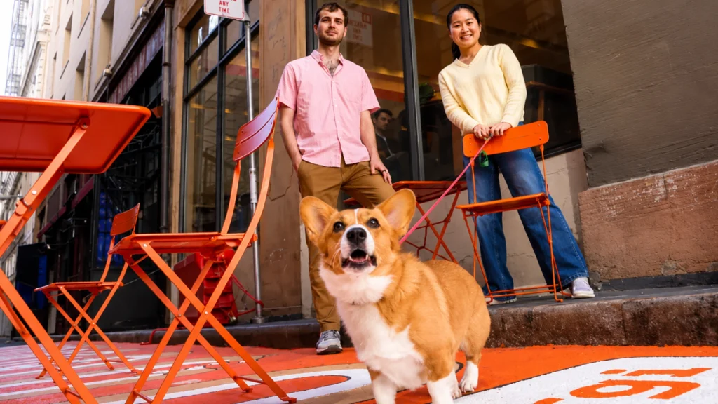 Corgi Insure Founders Nico Laqua and Emily Yuan with Corgi – Tumisang Bogwasi Nico Laqua and Emily Yuan standing outdoors with a corgi dog in front of orange chairs on a city street.
