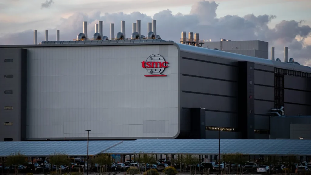 Large industrial facility with a curved white facade and a prominent red tsmc logo on the wall; a parking lot and landscaping visible in the foreground under a cloudy sky.