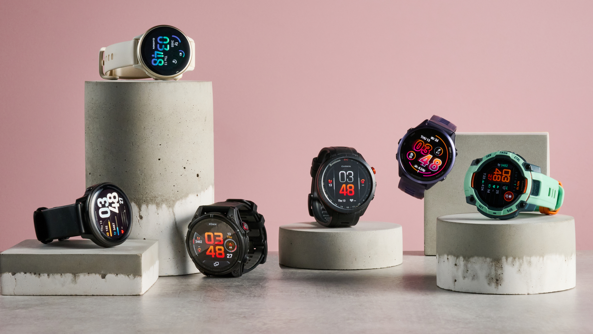 Multiple Garmin smartwatches displayed on concrete stands against a pink background, featuring different designs, colors, and digital watch faces.