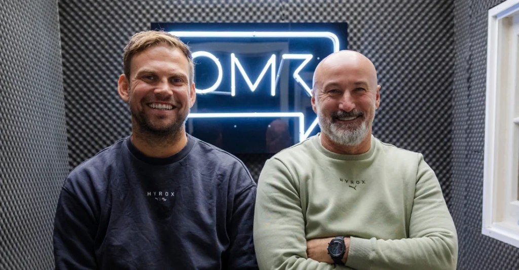 HYROX Founders Christian Toetzke and Moritz Fürste – Tumisang Bogwasi HYROX founders Christian Toetzke and Moritz Fürste smiling together in front of an illuminated OMR sign while wearing HYROX apparel.
