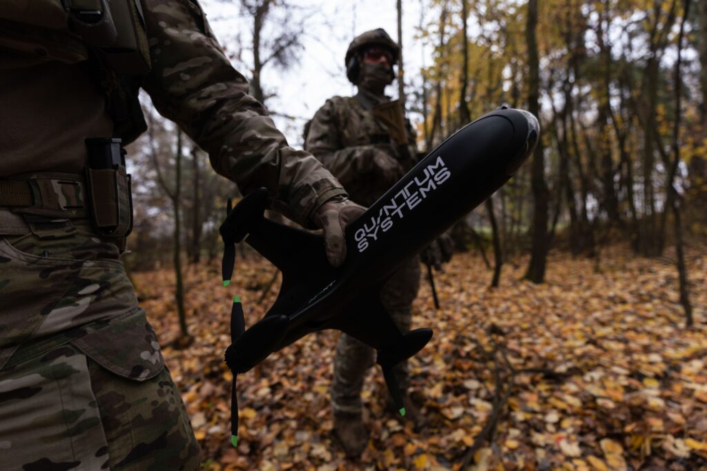 Quantum Systems Tactical Drone Supporting Field Operations – Tumisang Bogwasi Camouflaged soldier in a forest holds a large black cylindrical device labeled Quantum Systems, autumn leaves on the ground.