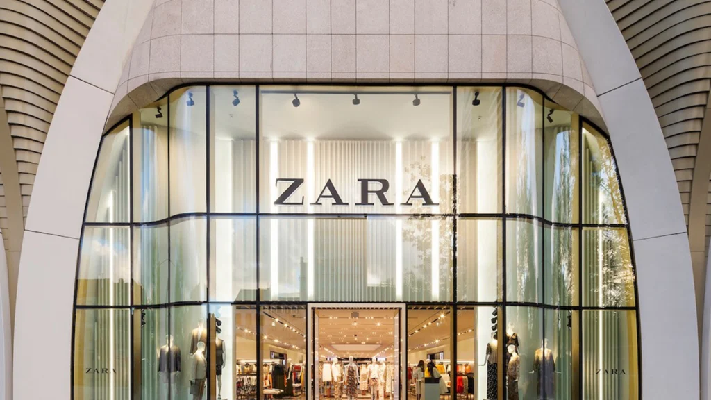 Zara Flagship Store with Modern Glass Façade – Tumisang Bogwasi Exterior of a Zara fashion store with a large curved glass façade, illuminated Zara signage, mannequins, and clothing displays visible inside.