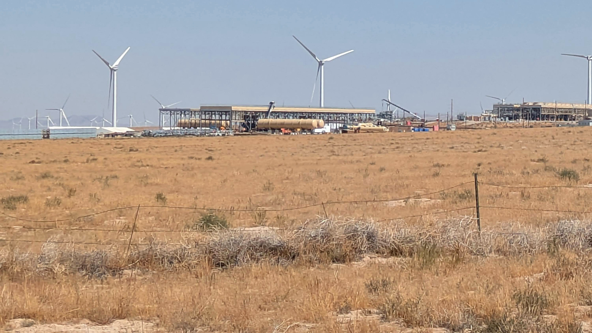 Fervo Energy's Cape Station geothermal plant under construction on open grassland in Utah, with wind turbines behind it