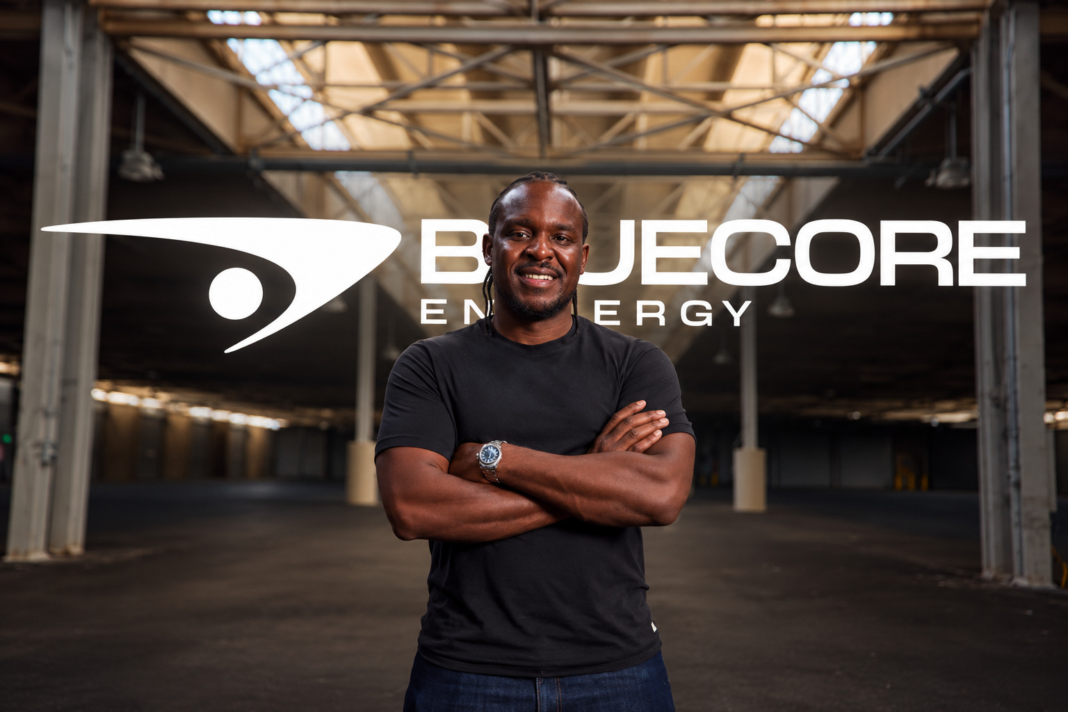 Kofi Asante, founder and CEO of Bluecore Energy, standing with arms folded in a warehouse in front of the Bluecore Energy logo